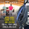 A coiled black cable secured with a Coil'n'Carry® Strap hangs on a hook by stage equipment as musicians set up instruments outdoors. Text: Audio, video, sound, stage, and lighting solutions for all your cords, cables, and gear.