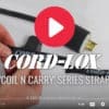 A person wraps a black Coil'n'Carry® Strap around an electrical cord near a large plug. The video displays the title COIL’N’CARRY® STRAP with a play button icon overlaid.