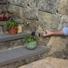 A hand reaches toward a modern doorbell mounted with VELCRO® Brand Industrial Strength Extreme Hook and Loop Strips (10 Pack) on a stone wall beside outdoor steps adorned with potted plants and flowers.