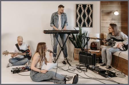 Four people are rehearsing as a band in a cozy room One plays keyboard one sings into a microphone and two play electric guitars There are music equipment cables and a plant in the background