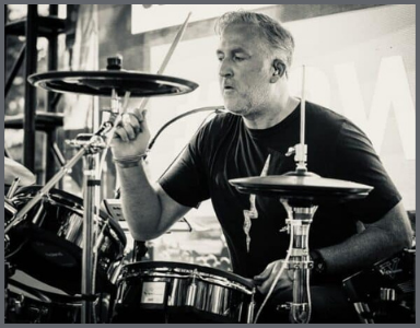 A man plays the drums passionately during a live performance He is holding drumsticks and wearing a dark T shirt The image is in black and white