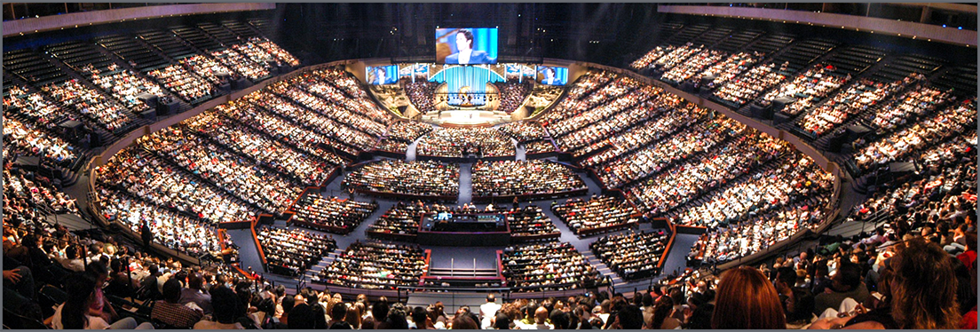 A large brightly lit auditorium filled with hundreds of seated people all facing a central stage with screens and speakers creating a sense of a major event or conference taking place