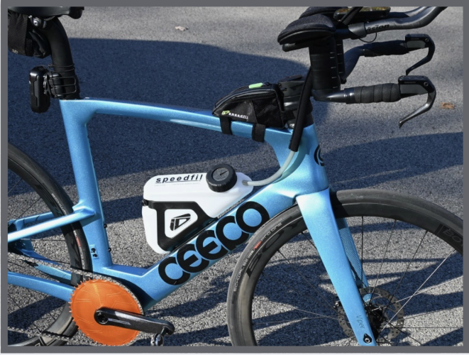 A light blue Ceepo triathlon bike with black handlebars a white Speedfil water bottle an orange chainring cover and a small black storage bag mounted on the top tube parked on gray pavement