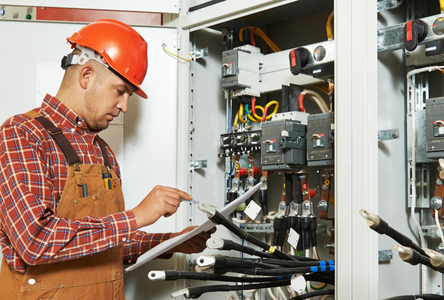 An electrician wearing a red hard hat and brown overalls checks a clipboard while inspecting electrical panels and cables inside an industrial control cabinet