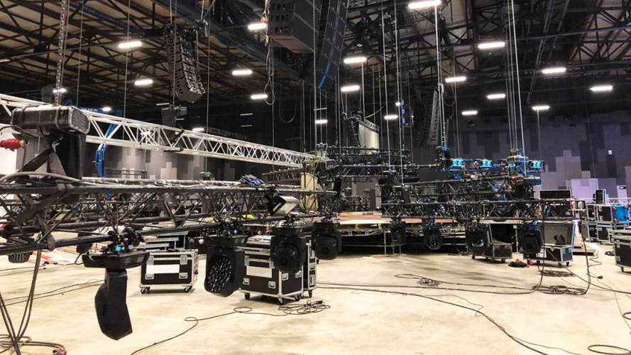 A large indoor venue with lighting rigs speakers metal trusses and cables set up for a stage production Equipment cases and wires are scattered across the concrete floor No people are visible