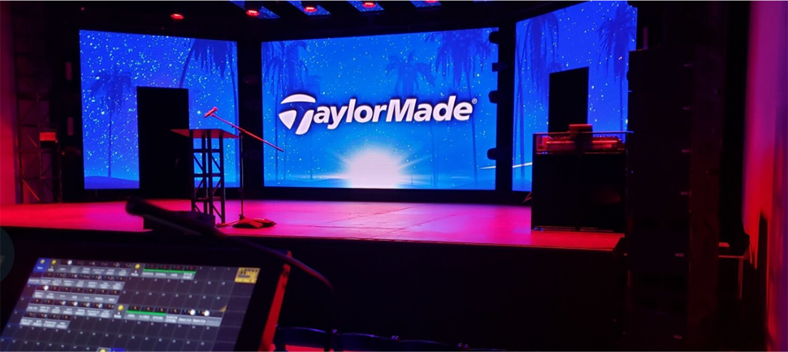 Stage setup for a TaylorMade presentation with a large blue screen backdrop showing the logo and palm trees pink lighting podium and audio equipment in front