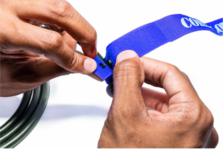 Closeup of two hands fastening a bright blue strap with metal rivets attaching it to a green cable against a white background TFI Custom Fabrication Close up of two hands fastening a bright blue strap with metal rivets attaching it to a green cable against a white background