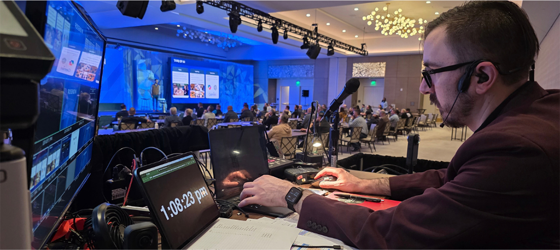 A man operates AV equipment at a control desk during a conference monitoring screens as attendees sit at tables in a large well lit event room with blue lighting and a stage in the background