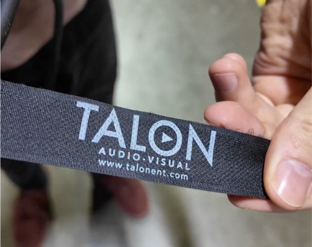 A hand holds a black strap with the text TALON AUDIO VISUAL wwwtalonentcom printed in white